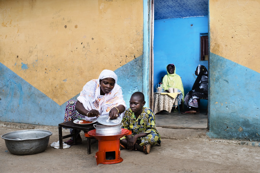 A woman and a boy are sitting in front of a house with a modern metal cookstove.