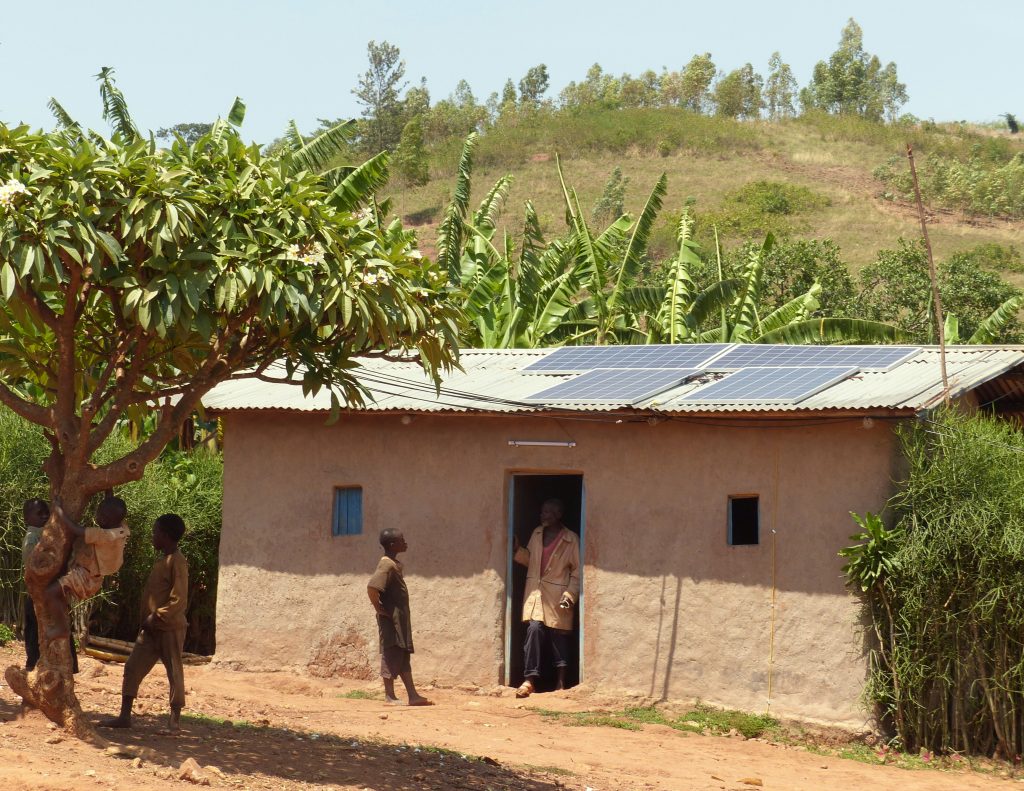 A Rwandan man is coming out of a small village house with solar panels on top while children are playing in a tree.