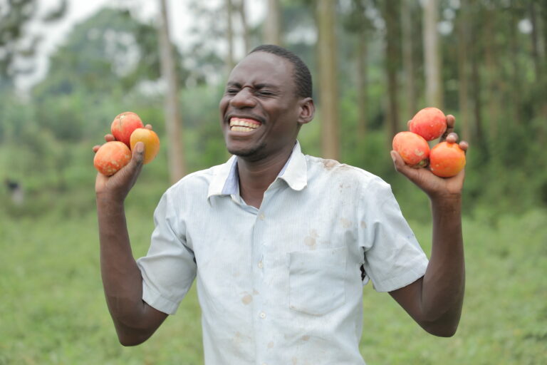 A happy looking man holding up three tomatos and each of his hands.