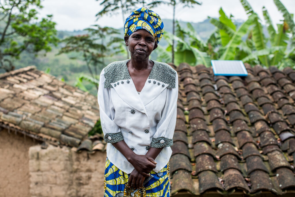 A female client standing in front of her home, where a Solar Home System has been installed on the roof.